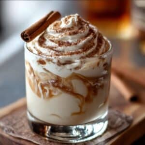 Creamy cinnamon roll cocktail with caramel swirls, whipped cream, and cinnamon dusting, topped with a cinnamon stick.