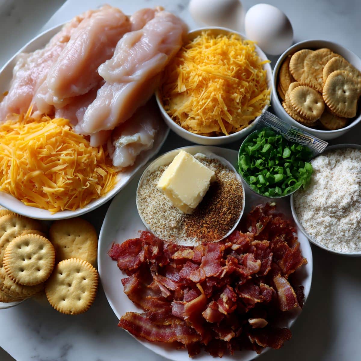 Ingredients for homemade crack chicken tenders including chicken, cream cheese, cheddar, bacon, ranch seasoning, and Ritz crumbs on a white counter.