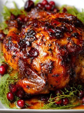 Cranberry maple roast chicken with glossy caramelized skin, fresh cranberries, and herbs on a serving platter.