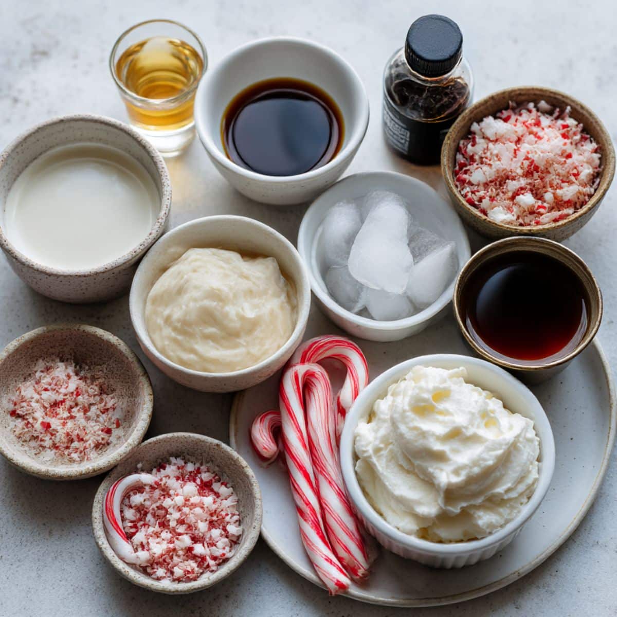 Peppermint white russian ingredients displayed in small bowls with crushed candy canes and cream on a white counter.