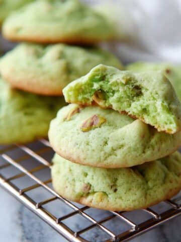 Soft pistachio pudding cookies stacked on a cooling rack with a bite showing their green, chewy centers.