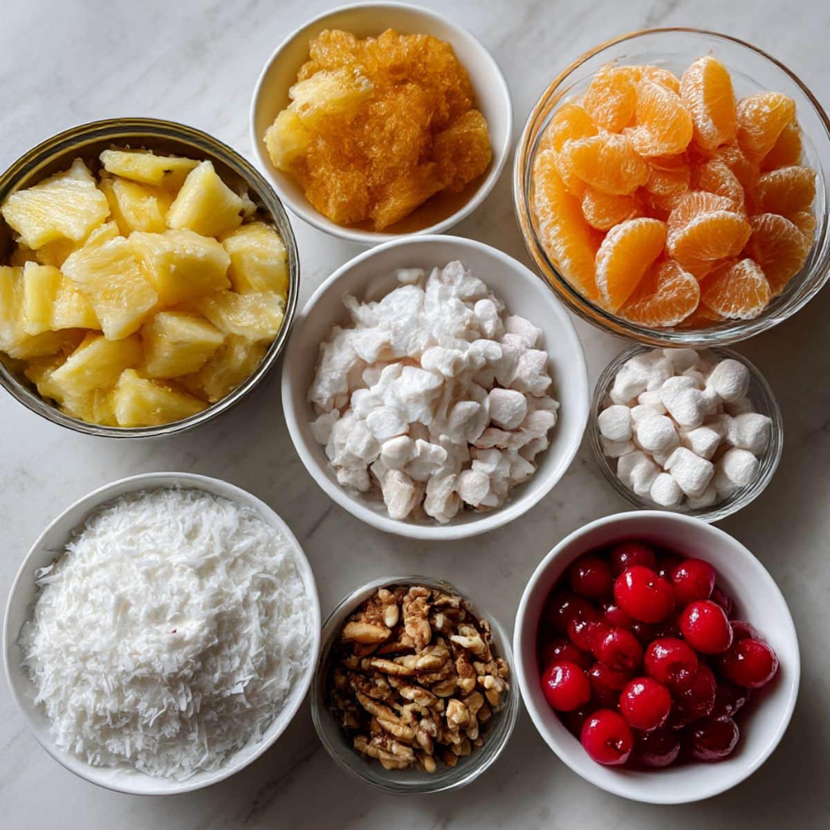 Ingredients for ambrosia fruit salad displayed on a white kitchen counter, including pineapple, oranges, coconut, marshmallows, and creamy mix-ins.