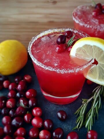 Cranberry Lemon Drop cocktail in a sugar-rimmed glass with ice, garnished with fresh cranberries, a lemon slice, and a rosemary sprig on a dark slate board.
