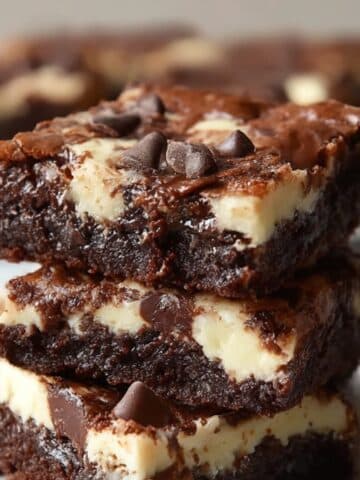 Rich, fudgy cream cheese brownies stacked on a plate, featuring a marbled swirl of chocolate and cheesecake layers with melted chocolate chips on top.