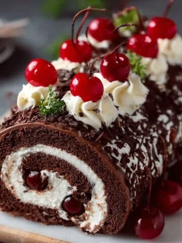 Elegant Black Forest cake roll topped with whipped cream rosettes, chocolate shavings, and bright cherries, sliced to show a chocolate sponge swirl with cherry-studded cream filling.
