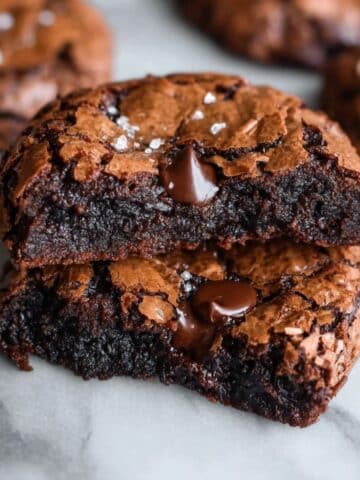 Fudgy chewy brookies stacked on a white marble counter, showing crackly brownie-like tops, gooey chocolate chips, and a dense fudgy center with flaky sea salt.
