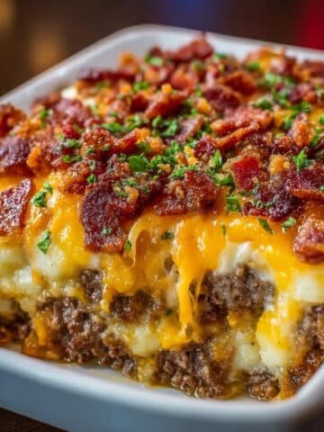 Garlic parmesan bacon cheeseburger casserole slice with melted cheddar, creamy layers, and crispy bacon topping in a white baking dish.
