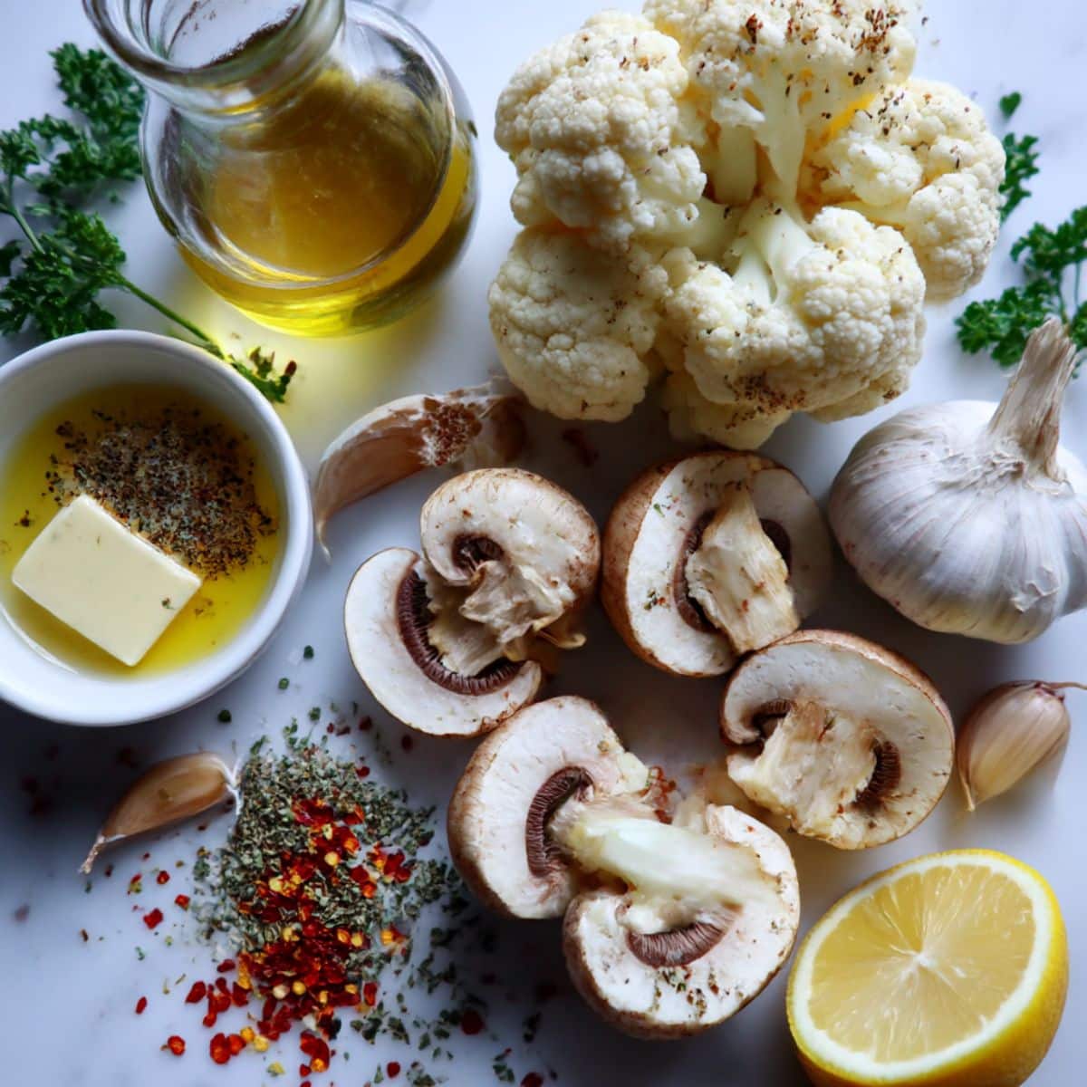 ngredients for garlic cauliflower and mushrooms, including cauliflower, mushrooms, garlic, olive oil, herbs, and lemon displayed on a white counter.