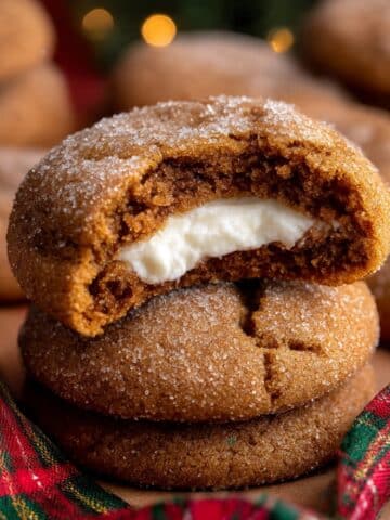 Stacked gingerbread cheesecake cookies filled with creamy cheesecake, with a bite taken out of the top cookie, resting beside festive ribbon on a parchment-lined surface.