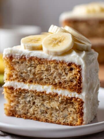 Moist, gluten free banana cake slice with creamy white frosting and banana slices on top.