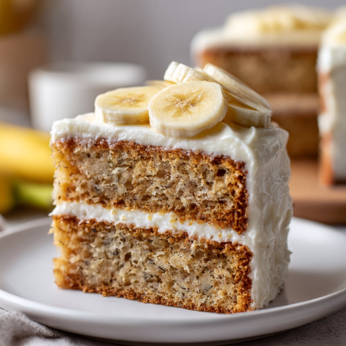 Moist, gluten free banana cake slice with creamy white frosting and banana slices on top.