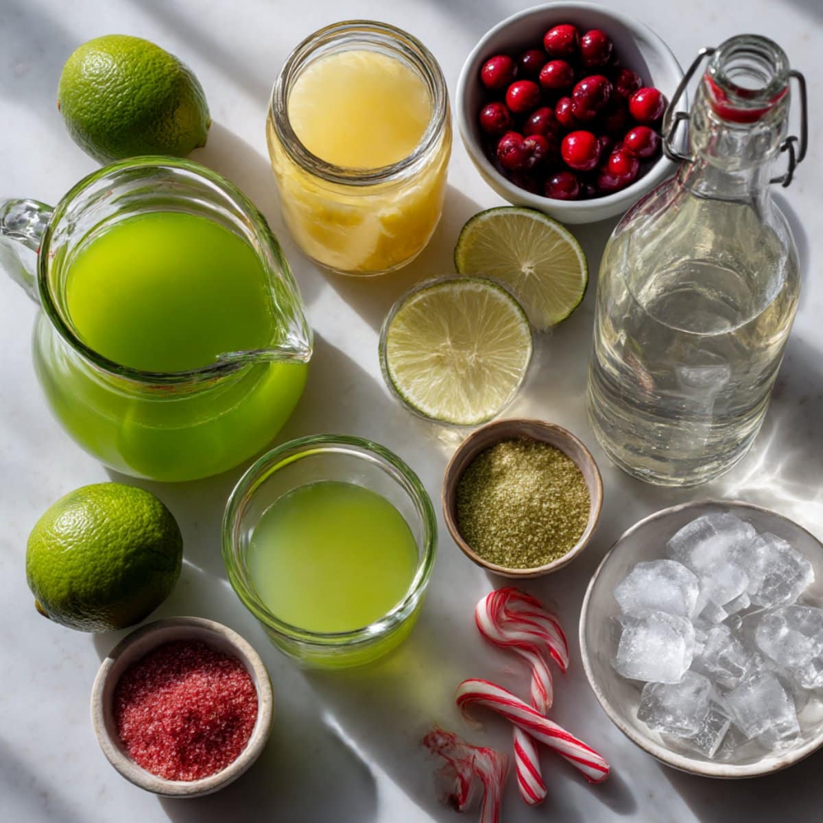 Grinch mimosa ingredients arranged on a white counter including green juice, pineapple juice, limes, and garnishes.