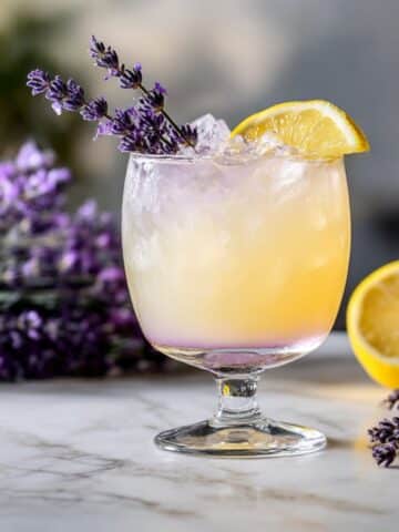 Lavender limoncello cocktail garnished with fresh lavender and lemon on a marble surface. A refreshing floral lemon drink with a light purple gradient.