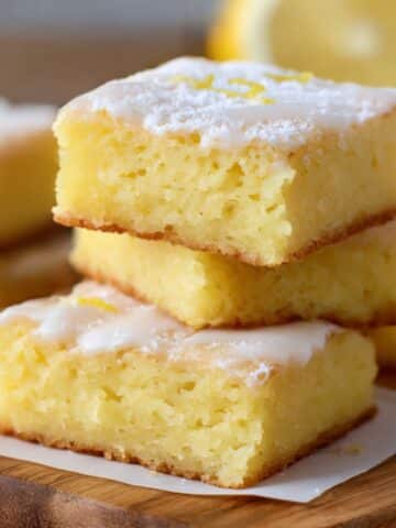 Lemon brownies with a soft, cakey crumb and glossy lemon glaze, stacked on a wooden board with fresh lemon zest on top.