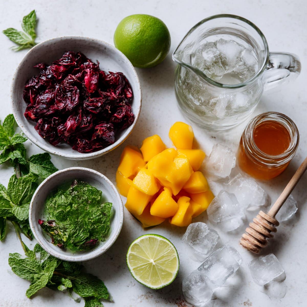 Ingredients for homemade mango hibiscus iced tea displayed on a white counter. Includes hibiscus, mango, lime, honey, mint, and ice for a fresh summer drink.