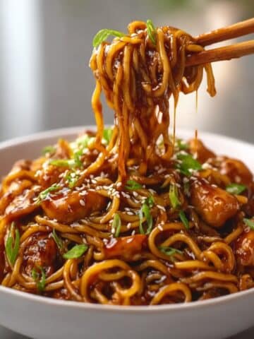 Garlic Chicken Noodles topped with sesame seeds and sliced green onions, with saucy noodles lifted by chopsticks from a white bowl.