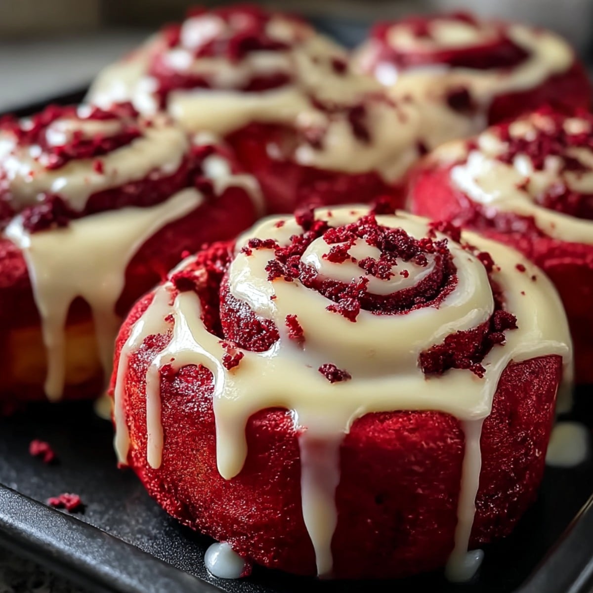 Red velvet cinnamon rolls in a baking pan, topped with thick cream cheese glaze dripping down the sides and sprinkled with red velvet crumbs.