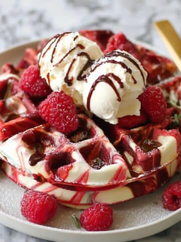 Red velvet marble waffles topped with vanilla ice cream, fresh raspberries, and chocolate drizzle on a white plate.