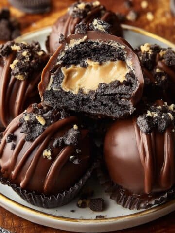 Reese’s Oreo Balls stacked on a plate, dipped in smooth chocolate with chocolate drizzle and Oreo crumbs, one cut open to show the creamy peanut butter center.