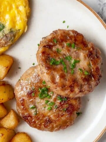 Turkey Breakfast Sausage patties on a white plate with crispy breakfast potatoes and omelet, sprinkled with chopped herbs.