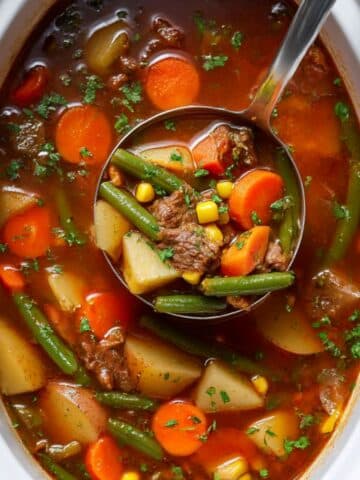 Vegetable beef soup in slow cooker, ladle lifting hearty mix of beef, potatoes, carrots, corn, and green beans in rich tomato broth with parsley garnish.