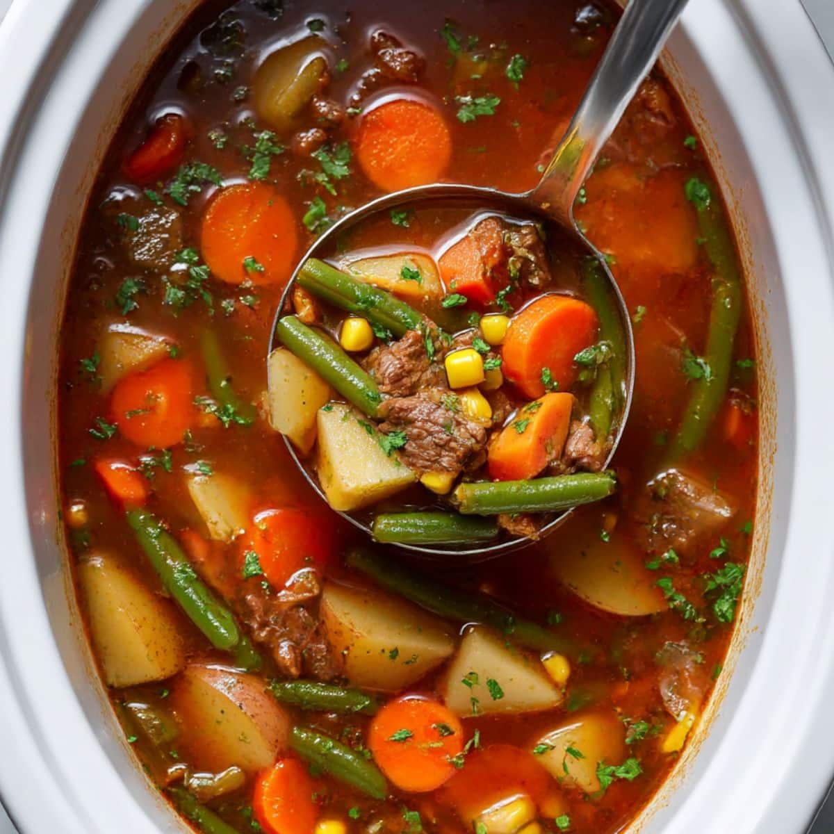 Vegetable beef soup in slow cooker, ladle lifting hearty mix of beef, potatoes, carrots, corn, and green beans in rich tomato broth with parsley garnish.