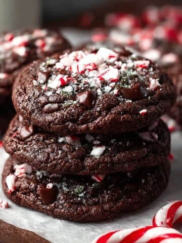 Double chocolate peppermint cookies stacked on parchment paper, topped with crushed candy canes and chocolate chips.