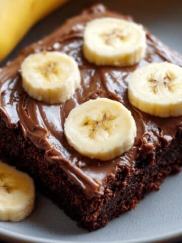 Banana brownie topped with creamy chocolate frosting and fresh banana slices, shown as a rich, fudgy square dessert on a plate.