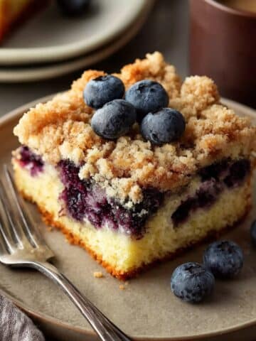 Moist blueberry coffee cake slice with a buttery crumb topping, layers of juicy blueberries, and fresh blueberries on top, served on a plate with a fork.