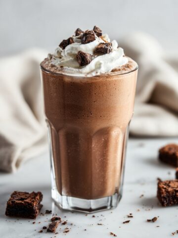 Tall glass of frozen brownie milkshake with creamy chocolate beverage, topped with swirls of whipped cream and chunks of fudgy brownies, served on a marble surface with brownie pieces scattered around and a white cloth napkin in the background.