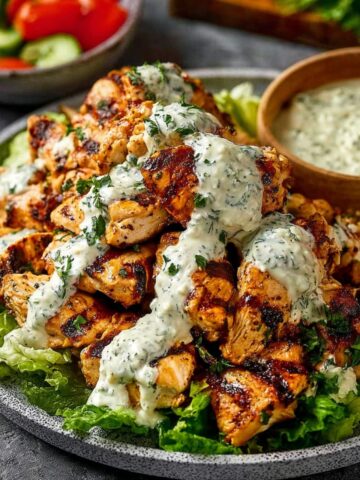 Grilled chicken shawarma with caramelized char marks served on lettuce, generously drizzled with creamy tahini sauce and fresh parsley, accompanied by a wooden bowl of extra sauce and a side salad with tomatoes and cucumbers