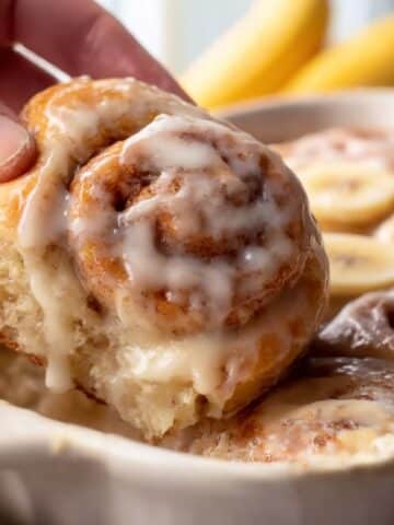 Banana bread cinnamon roll being pulled from a baking dish, glazed with rich vanilla icing and soft banana slices visible in the background.