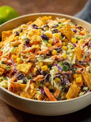 Cowboy Cabbage Salad served in a large white bowl on a wooden surface with fresh limes in the background, featuring shredded cabbage, carrots, corn kernels, black beans, crushed tortilla chips, and cilantro mixed in a creamy dressing