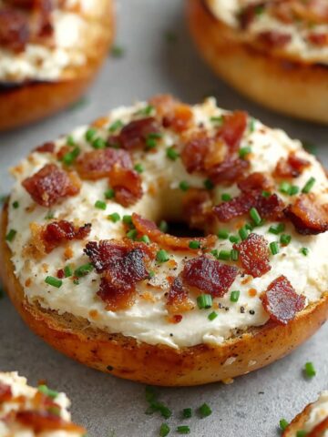 Toasted bagel topped with cream cheese, crispy bacon bits, fresh chives, and black pepper, with additional topped bagels in the background