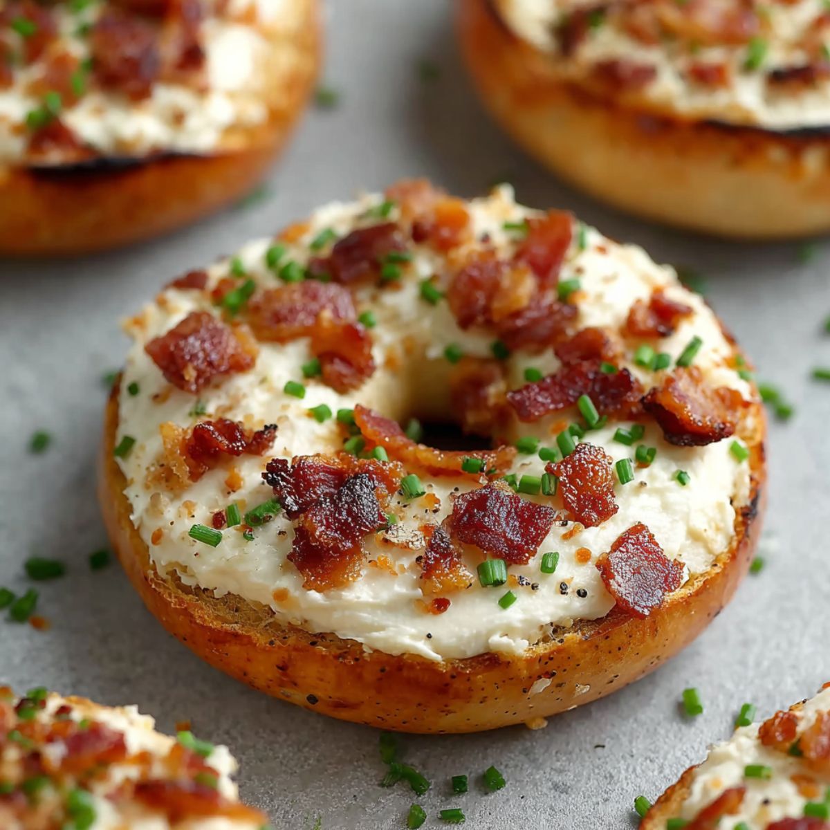 Toasted bagel topped with cream cheese, crispy bacon bits, fresh chives, and black pepper, with additional topped bagels in the background
