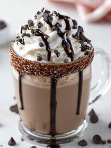 Dirty Snowman hot chocolate in a clear glass mug with a cinnamon-sugar rim, topped with fluffy whipped cream, chocolate drizzle, and mini chocolate chips.