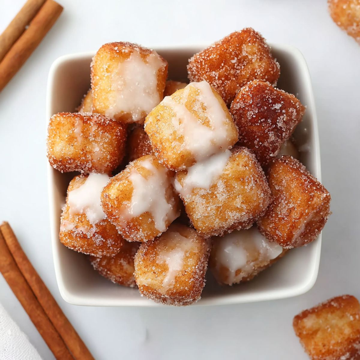 Angel cake churro bites coated in cinnamon sugar and drizzled with vanilla glaze, served in a white bowl with cinnamon sticks.