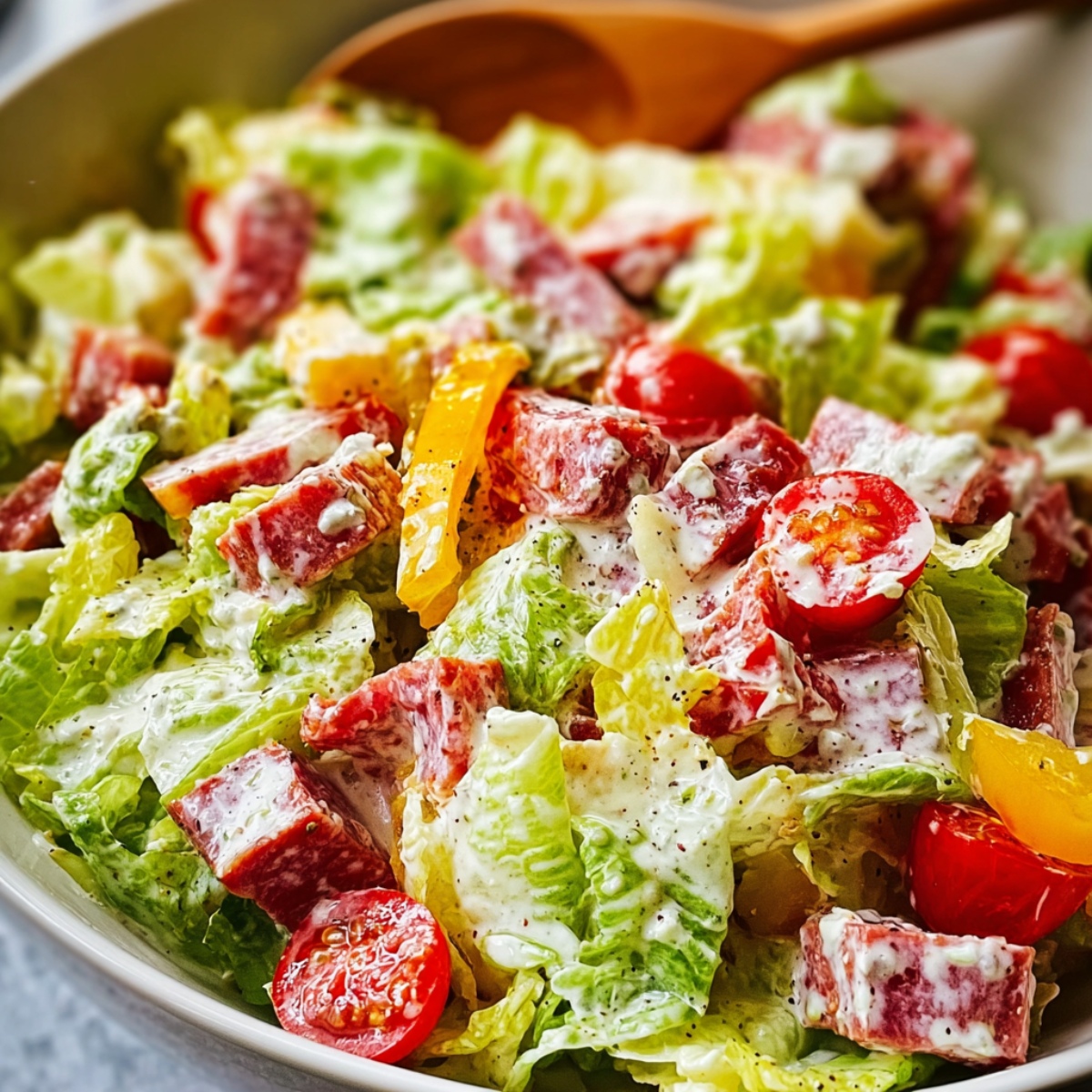 Easy Italian grinder salad with chopped romaine lettuce, salami, pepperoni, provolone cheese cubes, cherry tomatoes, and yellow bell pepper strips tossed in creamy Italian dressing with Parmesan and black pepper, served in a white bowl with wooden serving spoons