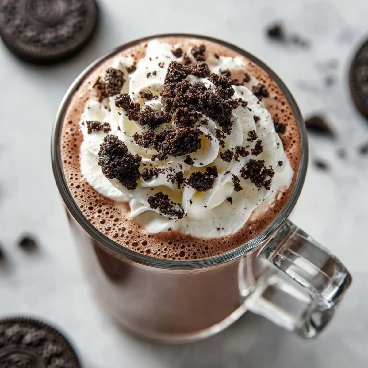Oreo hot chocolate in a clear glass mug, topped with whipped cream and crushed Oreo cookie pieces, photographed from an overhead angle.