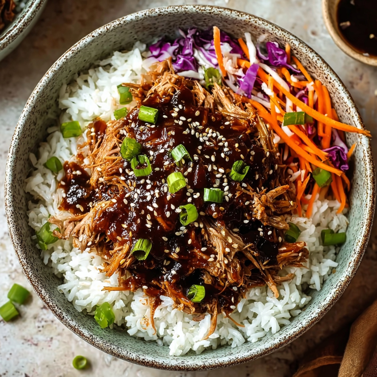 Easy Slow Cooker Asian Pulled Pork served in a speckled ceramic bowl over white rice, topped with tender shredded pork in dark Asian sauce, sesame seeds, and sliced green onions, accompanied by a colorful Asian slaw with shredded purple cabbage and julienned carrots