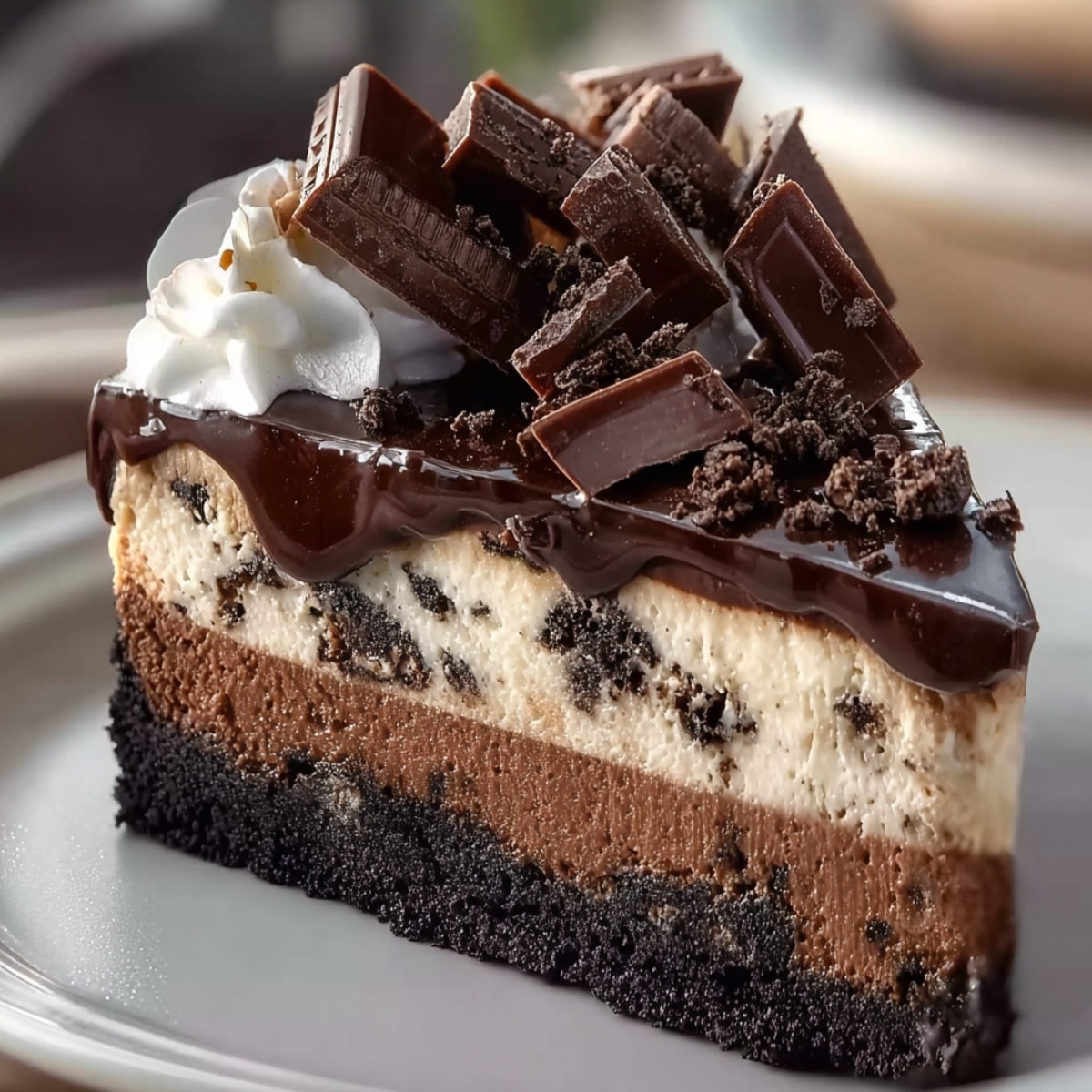 Triple chocolate cheesecake slice with Oreo cookie crust, creamy chocolate cheesecake layers, rich chocolate ganache topping, whipped cream, and chocolate chunks on a white plate.