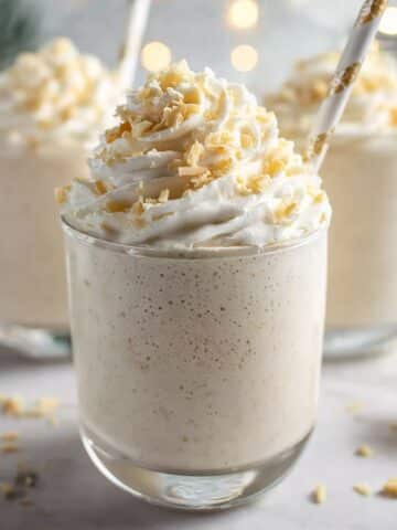 Easy winter wonderland smoothie in a clear glass, topped with fluffy whipped cream and white chocolate shavings, with festive holiday lights softly blurred in the background.
