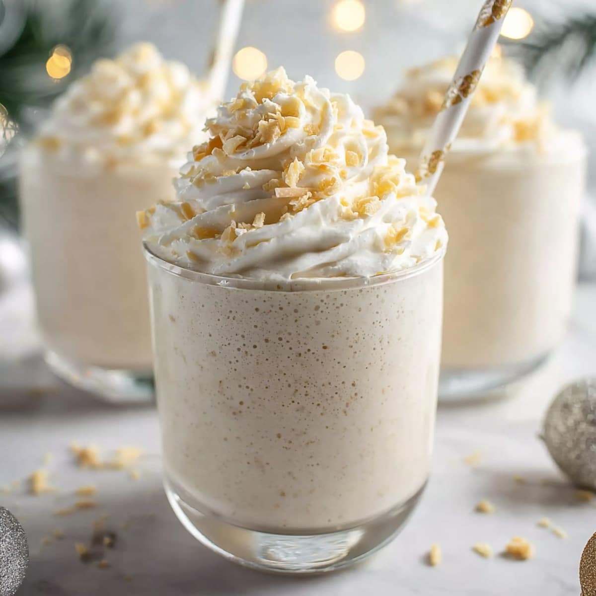 Easy winter wonderland smoothie in a clear glass, topped with fluffy whipped cream and white chocolate shavings, with festive holiday lights softly blurred in the background.