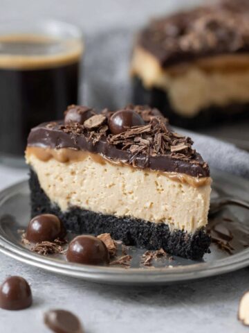 A slice of espresso cheesecake on a gray plate with a dark chocolate cookie crust, creamy tan espresso filling, and chocolate ganache topping garnished with chocolate shavings and chocolate-covered espresso beans, served alongside a cup of black coffee.