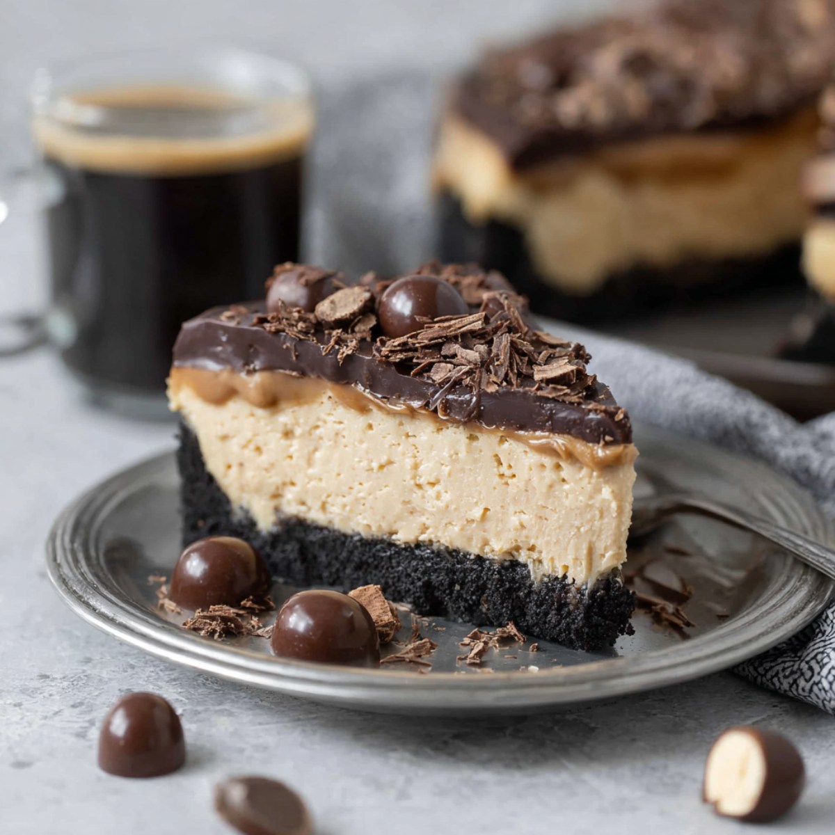 A slice of espresso cheesecake on a gray plate with a dark chocolate cookie crust, creamy tan espresso filling, and chocolate ganache topping garnished with chocolate shavings and chocolate-covered espresso beans, served alongside a cup of black coffee.