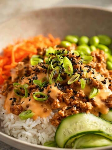 Ground turkey rice bowl topped with creamy sauce, sesame seeds, green onions, cucumber slices, shredded carrots, and edamame over fluffy white rice
