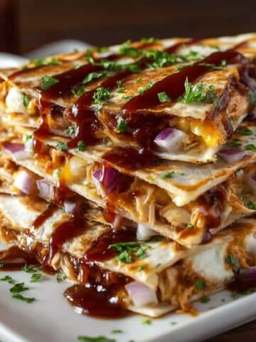 Hot honey BBQ chicken quesadillas stacked on a plate with shredded chicken, melted cheese, red onions, fresh herbs, and rich barbecue sauce drizzled on top