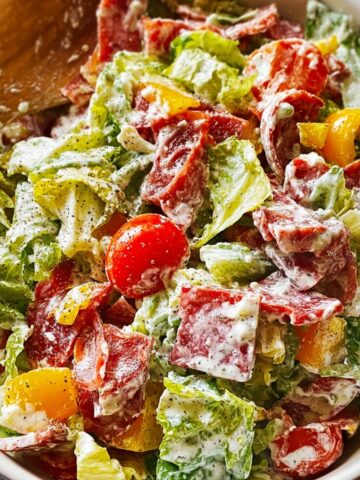Italian grinder salad in a white bowl with chopped romaine lettuce, rolled salami and pepperoni slices, cubed provolone cheese, cherry tomatoes, yellow bell peppers, and pepperoncini coated in creamy Italian dressing with grated Parmesan and cracked black pepper, served with wooden spoons