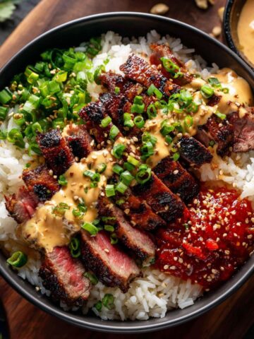 Korean BBQ steak rice bowl with sliced grilled steak showing char marks over white rice, drizzled with creamy yum yum sauce, topped with sliced green onions, sesame seeds, and gochujang (Korean red chili paste) on the side