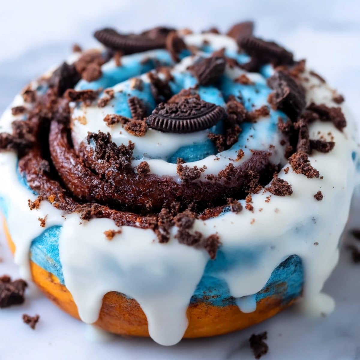 Cookie Monster cinnamon roll topped with bright blue frosting, white glaze drizzle, and crushed chocolate sandwich cookies.